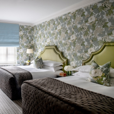 Hotel family room with two Queen-sized beds, each featuring a green upholstered headboard. The walls are covered in green and white floral wallpaper, with matching decorative pillows on the beds.